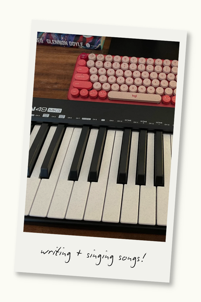 Pink typing keyboard and a piano MIDI keyboard on a desk. A Polaroid frame surrounds the photo, and text on the frame reads "writing and singing songs!"