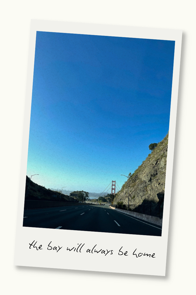 The Golden Gate Bridge from a distance, with a hill on the right. A Polaroid frame surrounds the photo, and text on the frame reads "the bay will always be home."